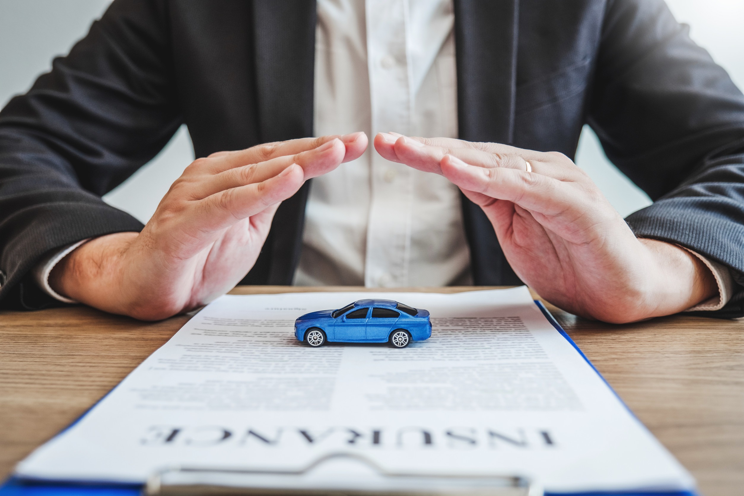 A person in a suit holds their hands protectively over a small blue toy car placed on an insurance document, symbolizing Minnesota auto insurance coverage and protection.