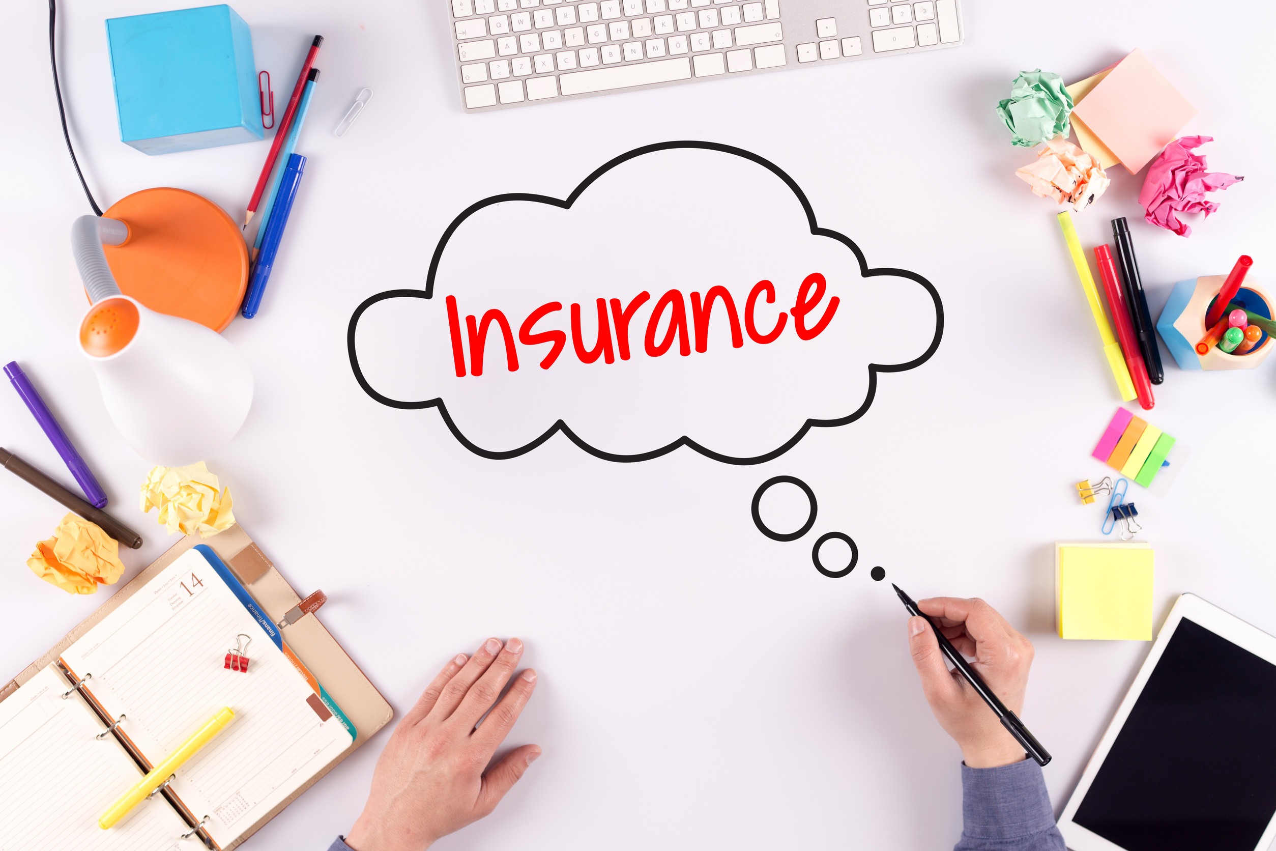 A person’s hands at a cluttered desk, with colorful stationery and an open planner, drawing a thought bubble containing the word “Insurance” in red on a white sheet of paper.