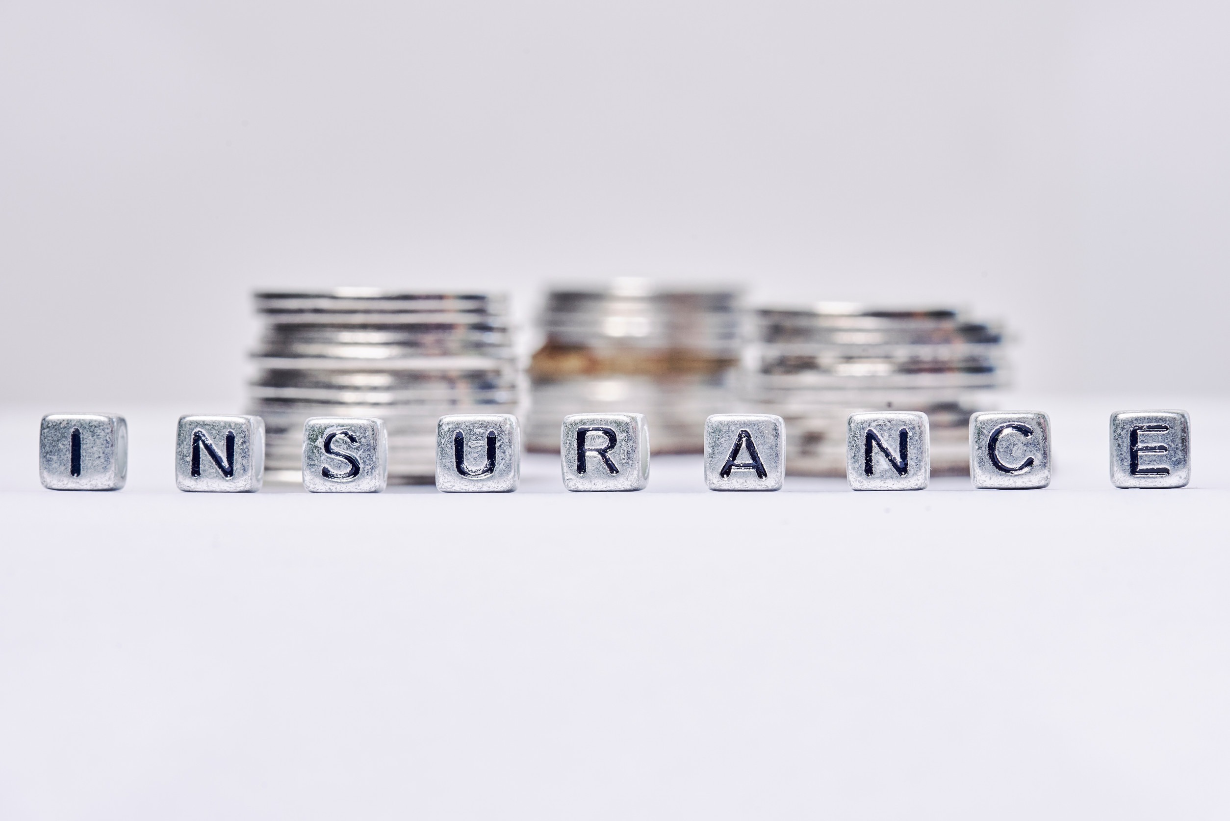 Silver letter blocks spelling “INSURANCE” are lined up in front of blurry stacks of coins on a white surface, symbolizing the range of coverage options available through independent insurance providers in Minnesota.