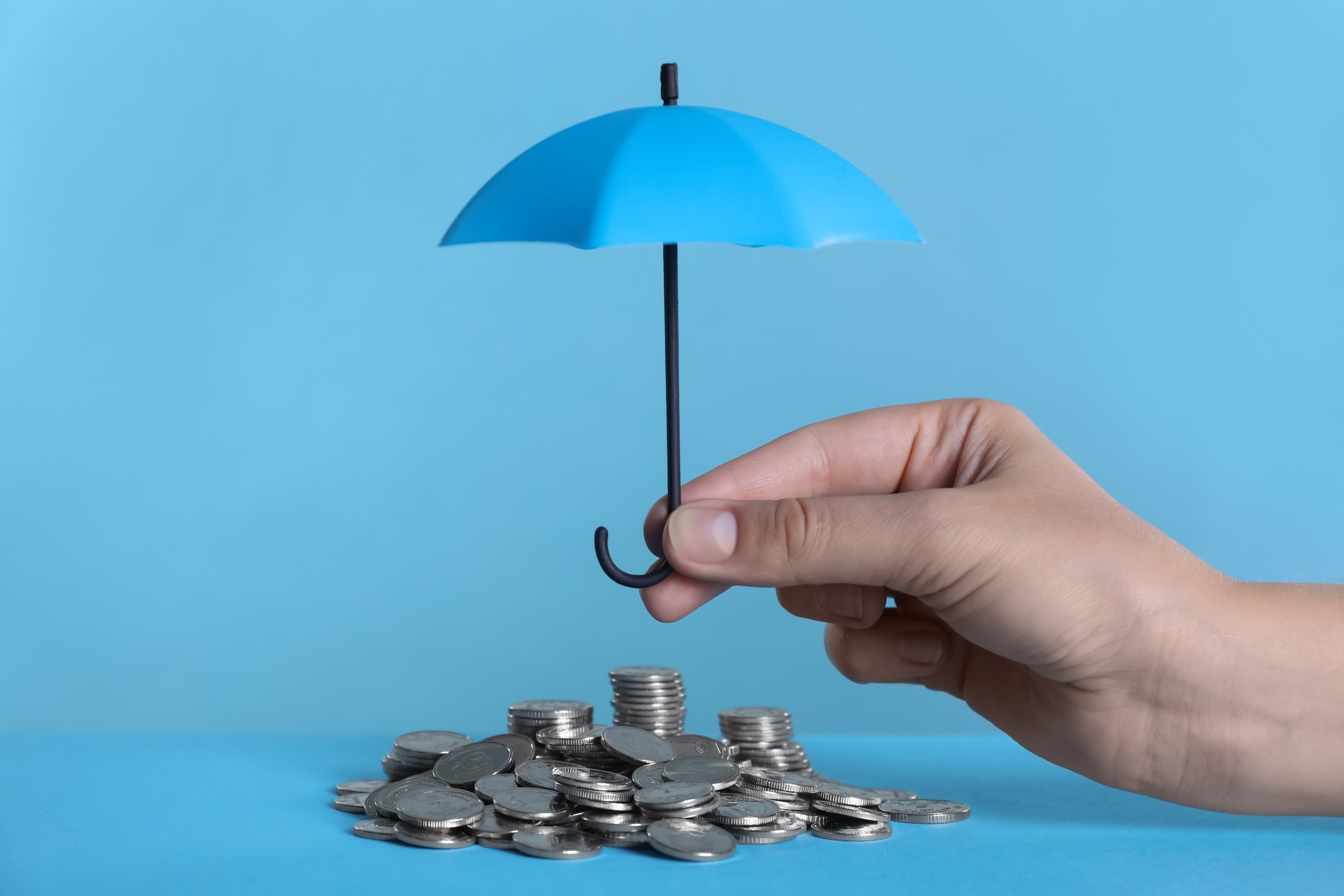 A hand holds a small blue umbrella over stacks and piles of coins, symbolizing financial protection or savings insurance, against a blue background.